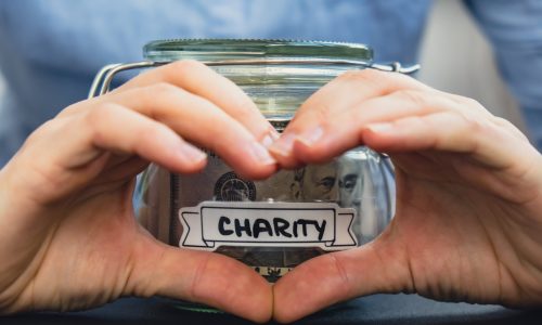 Female showing heart sign with hands Saving Money In Glass Jar filled with Dollars banknotes. CHARITY transcription in front of jar. Managing personal finances extra income for future insecurity background